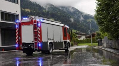 Rotes Feuerwehrfahrzeug mit leuchtenden blauen Warnlichtern parkt auf nasser Straße in bergiger Landschaft.