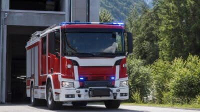 Rotes und weißes Feuerwehrfahrzeug mit blauen Blaulichtern vor einer Feuerwehrhalle in bergiger Landschaft