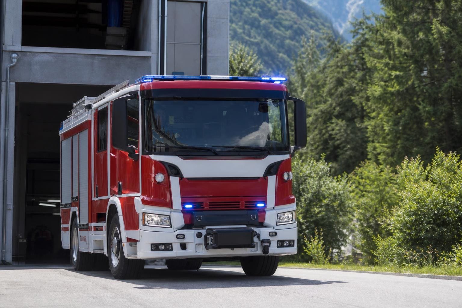 Rotes und weißes Feuerwehrfahrzeug mit blauen Blaulichtern vor einer Feuerwehrhalle in bergiger Landschaft