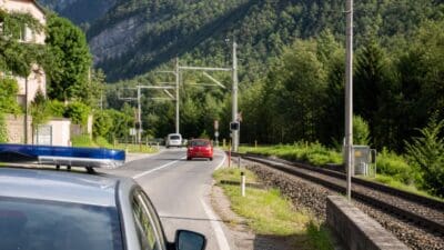 Polizeifahrzeug mit eingeschaltetem Blaulicht auf einer Landstraße neben Bahngleisen vor einer Bergkulisse.