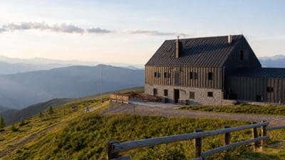 Ein dunkles Holzhaus mit Steinfundament steht auf einer grünen Bergwiese vor einem Bergpanorama.