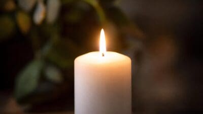 Eine brennende weiße Kerze mit heller Flamme vor einem unscharfen grünen Hintergrund.