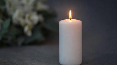 Eine brennende weiße Kerze auf einer dunklen Fläche vor unscharfen weißen Blumen.