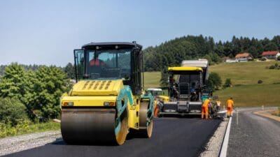 Gelbe und türkise Straßenwalze verdichtet frischen Asphalt auf einer Landstraße mit Bauarbeitern im Hintergrund