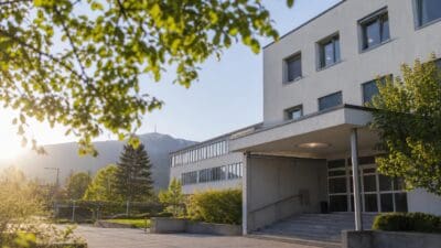 Modernes graues Gebäude mit vielen Fenstern und Eingangsbereich vor einer Bergkulisse