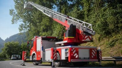 Rotes Feuerwehrfahrzeug mit ausgefahrener Drehleiter auf einer kurvigen Straße vor grünen Bäumen.