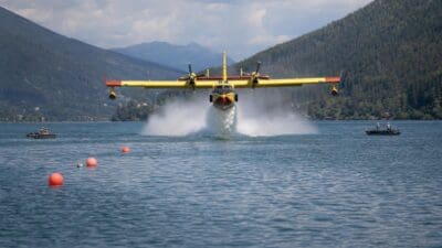 Gelbes Löschflugzeug mit zwei Propellern fliegt über dem blauen See und erzeugt eine große Wasserfontäne.