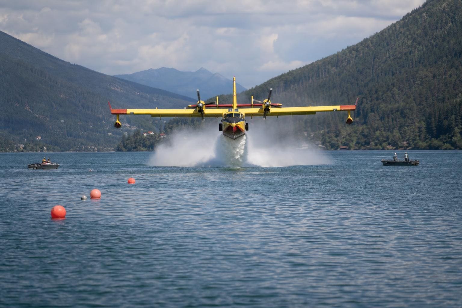 Gelbes Löschflugzeug mit zwei Propellern fliegt über dem blauen See und erzeugt eine große Wasserfontäne.