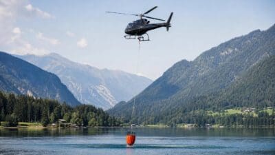 Schwarzer Hubschrauber mit rotem Behälter entnimmt Wasser aus dem Weissensee