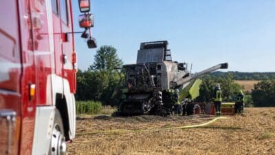 Rotes Feuerwehrauto im Vordergrund neben einem beschädigten Mähdrescher mit Feuerwehrleuten auf einem Feld