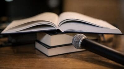 Ein aufgeschlagenes Buch liegt auf zwei geschlossenen Büchern, davor steht ein Mikrofon auf einem Holztisch.