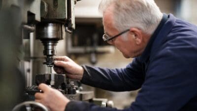 Ein älterer Mann mit weißem Haar und Brille bedient eine Werkzeugmaschine in der Werkstatt.