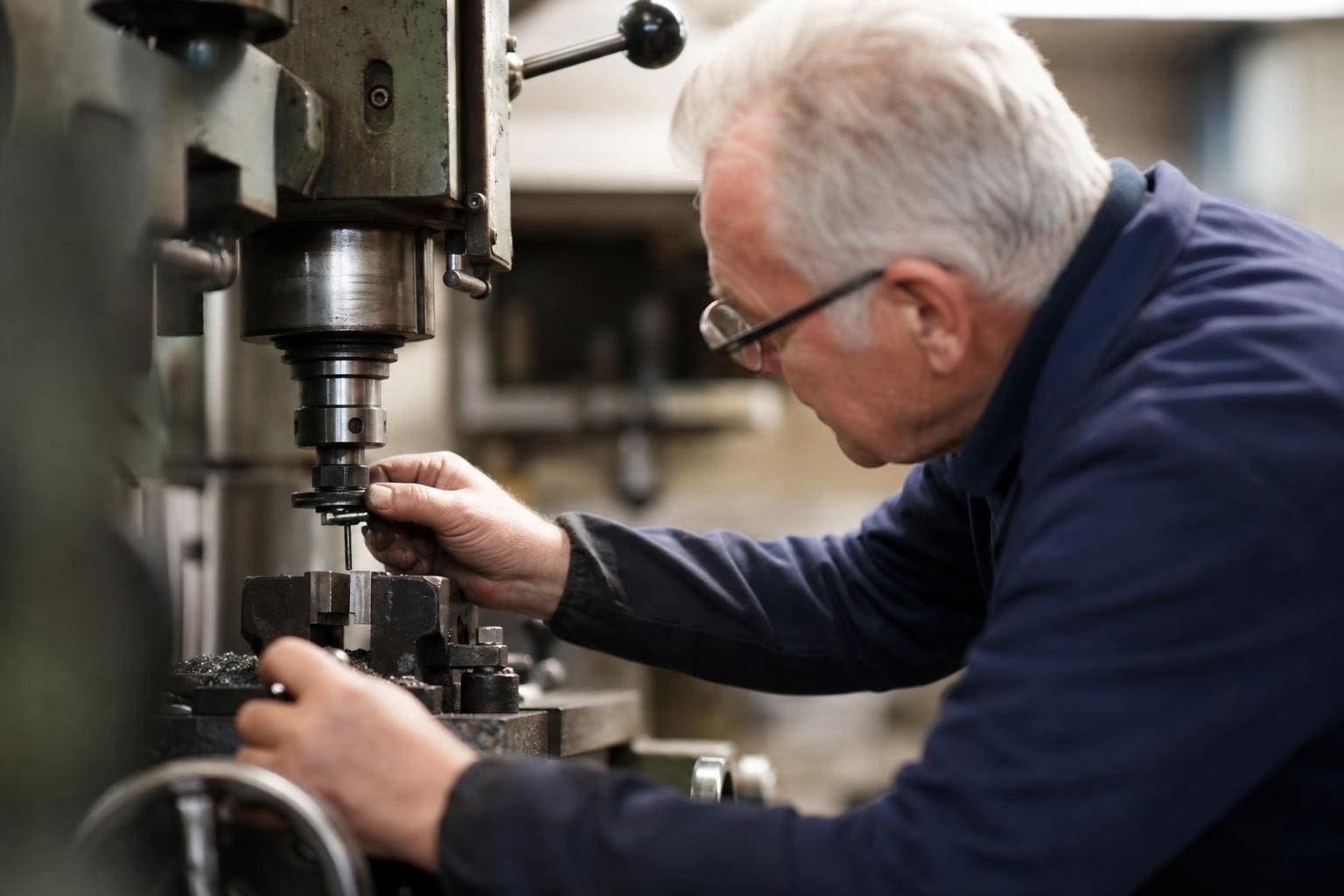 Ein älterer Mann mit weißem Haar und Brille bedient eine Werkzeugmaschine in der Werkstatt.