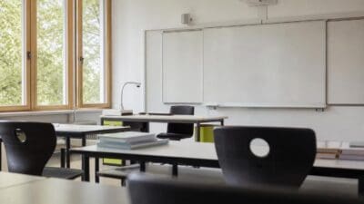 Leeres Klassenzimmer mit Schultischen, Stühlen und großen weißen Tafeln vor Fenstern mit Blick auf Bäume.