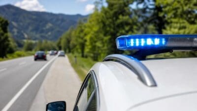 Weißes Polizeifahrzeug mit blauem Blaulichtbalken auf einer Landstraße vor Bergen und Bäumen