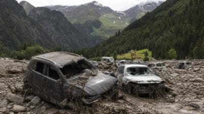 Mehrere stark beschädigte und schlammbedeckte Autos in einer Gerölllandschaft vor Bergen
