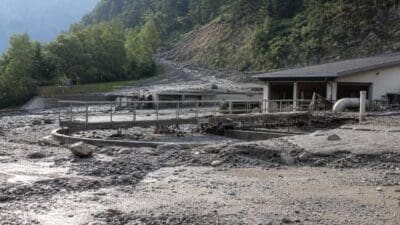 Die von Geröll und Schlamm bedeckte Kläranlage Sölden mit umliegenden Gebäuden und Wald im Hintergrund