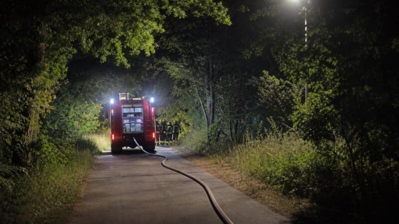Rotes Feuerwehrfahrzeug parkt nachts auf einem Waldweg mit Feuerwehrleuten und Schlauch auf dem Boden.