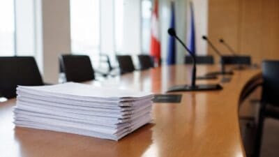 Ein Stapel weißer Papiere liegt auf einem Holztisch in einem Konferenzraum mit Mikrofonen und Stühlen.