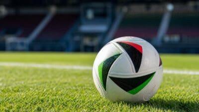 Ein weißer Fußball mit grünen schwarzen und roten Mustern liegt auf grünem Rasen in einem Stadion.