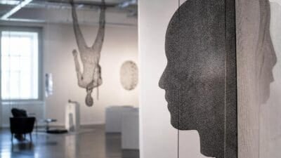 Drahtskulptur eines Kopfprofils und einer hängenden Figur in einer hellen Ausstellungshalle.