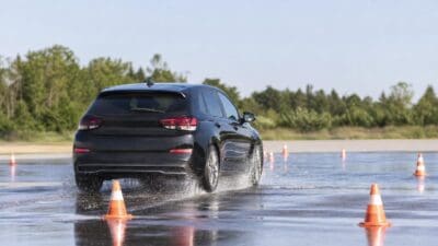 Ein schwarzer Kombi fährt durch eine Wasserlache auf einer Teststrecke mit orangen Verkehrskegeln.