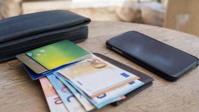 Kreditkarten, Euro-Banknoten und ein Smartphone liegen auf einem Holztisch neben Geldbörsen.