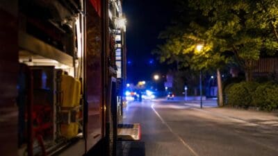 Seitenansicht eines roten Feuerwehrfahrzeugs auf einer nächtlichen Straße mit Bäumen und Laternen