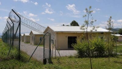 Stacheldrahtzaun mit offenem Tor vor gelben Gebäuden in ländlicher Umgebung unter blauem Himmel