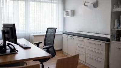 Arztpraxis mit Schreibtisch, Bürostuhl, Computer und Untersuchungsliege in einem hellen Raum