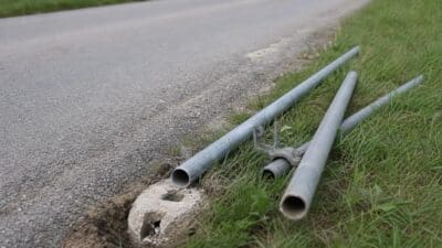 Graue Metallrohre und zerstörtes Betonfundament liegen am grasbewachsenen Straßenrand neben einer Asphaltstraße.