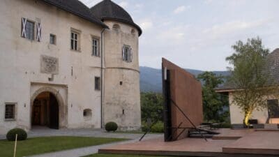 Historisches Schloss mit runder Turm und moderner Freilichtbühne mit großer brauner Wand im Vordergrund.