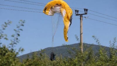 Gelber Paragleitschirm verfangen in Stromleitungen an einem Mast mit hängendem Gurtzeug vor bewaldeten Hügeln