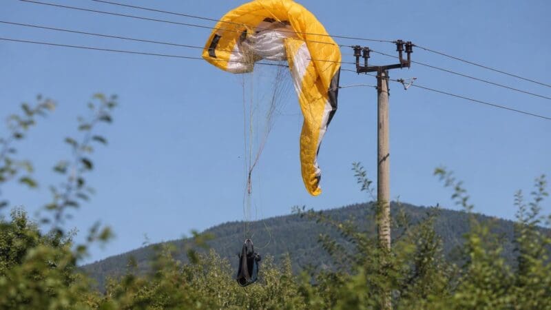 Gelber Paragleitschirm verfangen in Stromleitungen an einem Mast mit hängendem Gurtzeug vor bewaldeten Hügeln