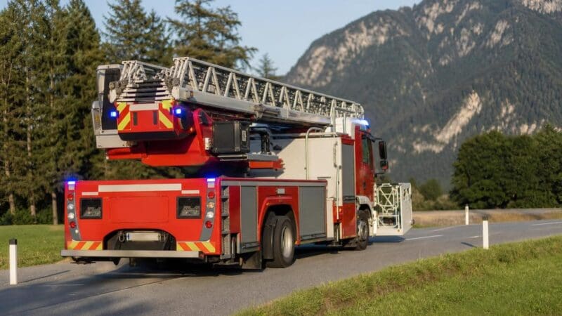 Rotes Feuerwehrfahrzeug mit Drehleiter und Blaulicht auf einer Landstraße vor Bergen