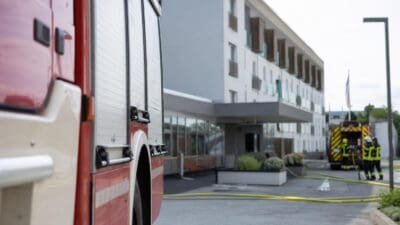 Rotes Feuerwehrauto im Vordergrund, dahinter ein Gebäude mit Feuerwehrleuten und gelben Schläuchen auf dem Platz.