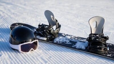 Ein schwarzer Snowboardhelm mit Skibrille liegt neben einem Snowboard im Schnee.