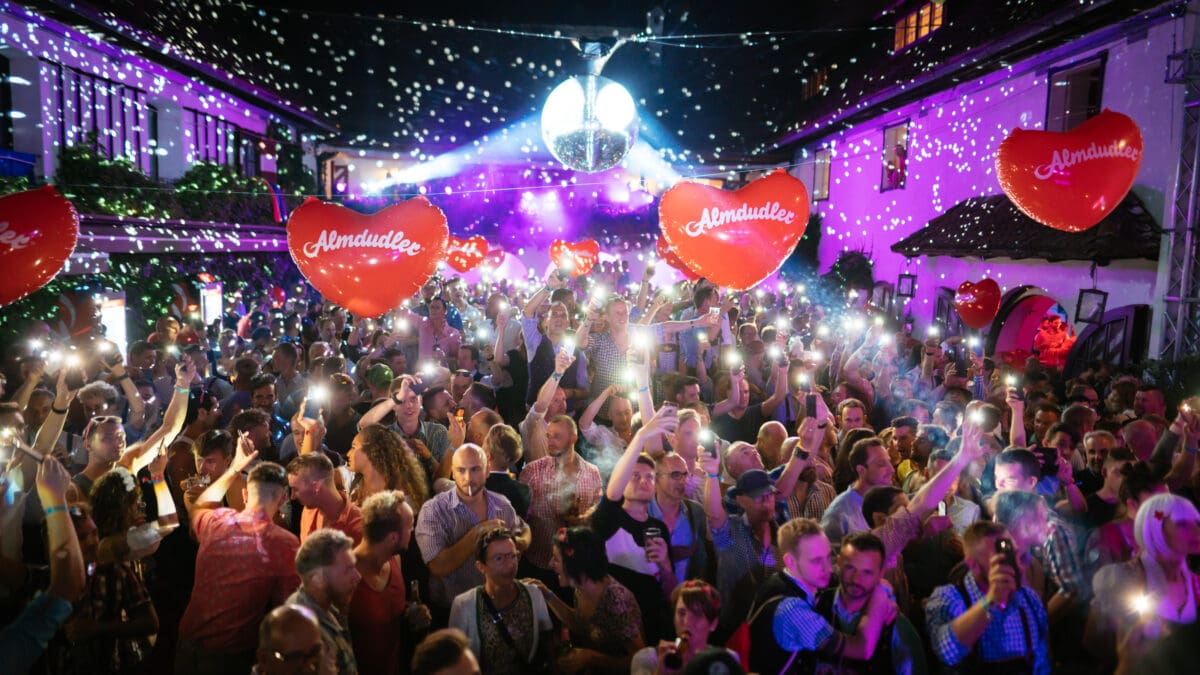 Dichte Menschenmenge bei einer nächtlichen Veranstaltung mit Disco-Kugel und roten Almdudler-Herzballons.