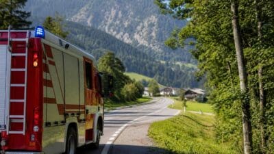 Rotes und weißes Feuerwehrfahrzeug parkt am Rand einer kurvigen Landstraße vor Bergen.