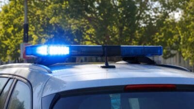 Blaues Blaulicht leuchtet auf dem Dach eines silbernen Polizeifahrzeugs vor grünen Bäumen.