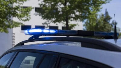 Blaues Blaulicht leuchtet auf dem Dach eines Polizeifahrzeugs vor grünen Bäumen und einem Gebäude.