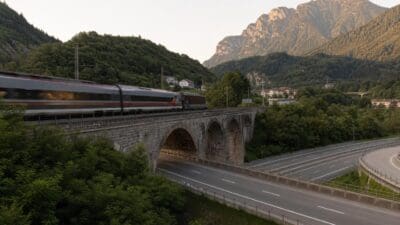 Ein Zug fährt über eine steinerne Bogenbrücke über einer Autobahn in einer bergigen Landschaft.