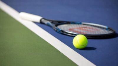 Ein gelber Tennisball liegt vor einem Tennisschläger auf einem blauen Tennisplatz.