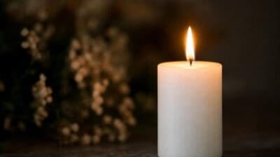 Eine brennende weiße Kerze steht auf einer dunklen Oberfläche vor unscharfen Blumen.