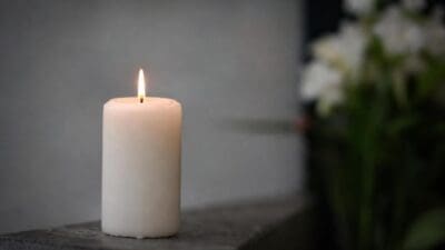 Eine brennende weiße Kerze auf einer dunklen Steinplatte mit unscharfen weißen Blumen im Hintergrund