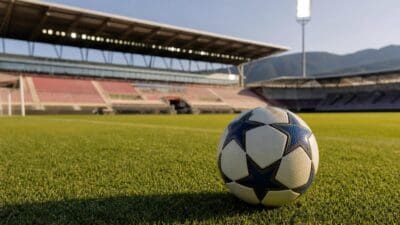Ein Fußball mit Sternenmuster liegt auf dem grünen Rasen eines leeren Stadions mit Tribünen und Bergen im Hintergrund.