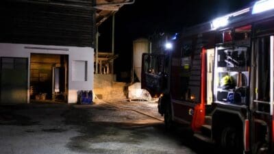 Rotes Feuerwehrfahrzeug parkt nachts vor einem Gebäude mit offen stehender Tür und Schlauch auf dem Boden