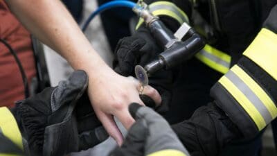 Feuerwehrleute in schwarzen Handschuhen mit gelben Reflexstreifen bearbeiten mit einem rotierenden Trennwerkzeug den Ring an einem Finger.