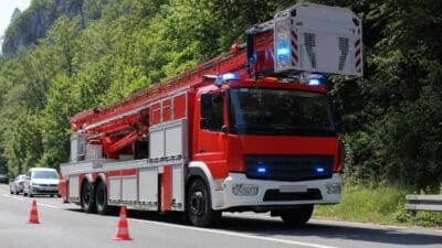 Rotes Feuerwehrfahrzeug mit Drehleiter und Blaulicht auf einer Straße vor bewaldetem Hang