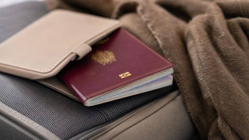 Ein roter Reisepass mit goldenem Emblem steckt in einer beigen Hülle auf einem Koffer.